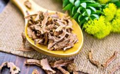 Auf einem Holztisch befindet sich eine Bastmatte mit Rhodiola-Kraut und Blüten und ein Löffel mit Rhodiola Wurzel