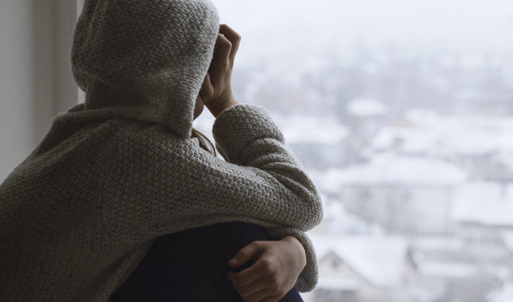 Mädchen sitzt niedergeschlagen am Fenster und schaut traurig nach draußen auf die verschneite, graue Stadt