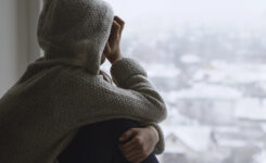 Mädchen sitzt niedergeschlagen am Fenster und schaut traurig nach draußen auf die verschneite, graue Stadt