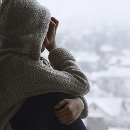 Mädchen sitzt niedergeschlagen am Fenster und schaut traurig nach draußen auf die verschneite, graue Stadt